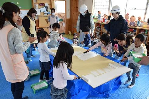 展示用パネルに道具の使い方、塗り方の指導をうけている園児とそれを写真に摂っている先生方の写真