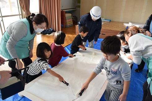園児が展示用パネルに色を塗って先生方がそれをみている写真
