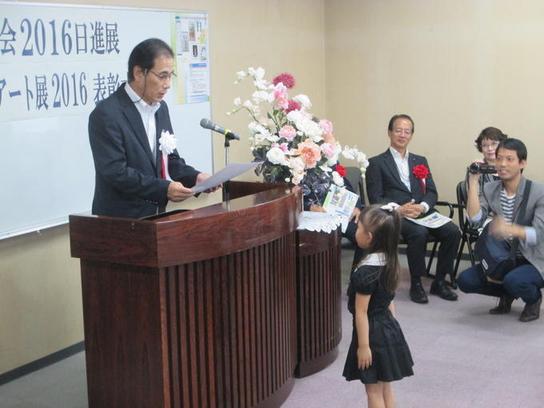 日進市民美術展覧会「2016日進展」とにっしん子どもアート展2016の表彰式にて女児に表彰状を読み上げる男性の写真