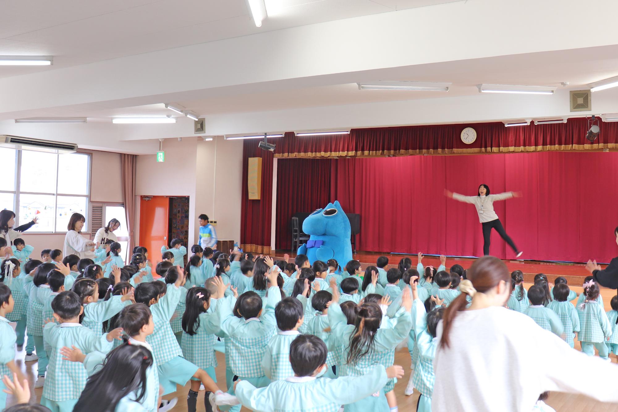 ステージ上の先生に倣ってにっしん体操を踊るニッシーとこどもたちの写真