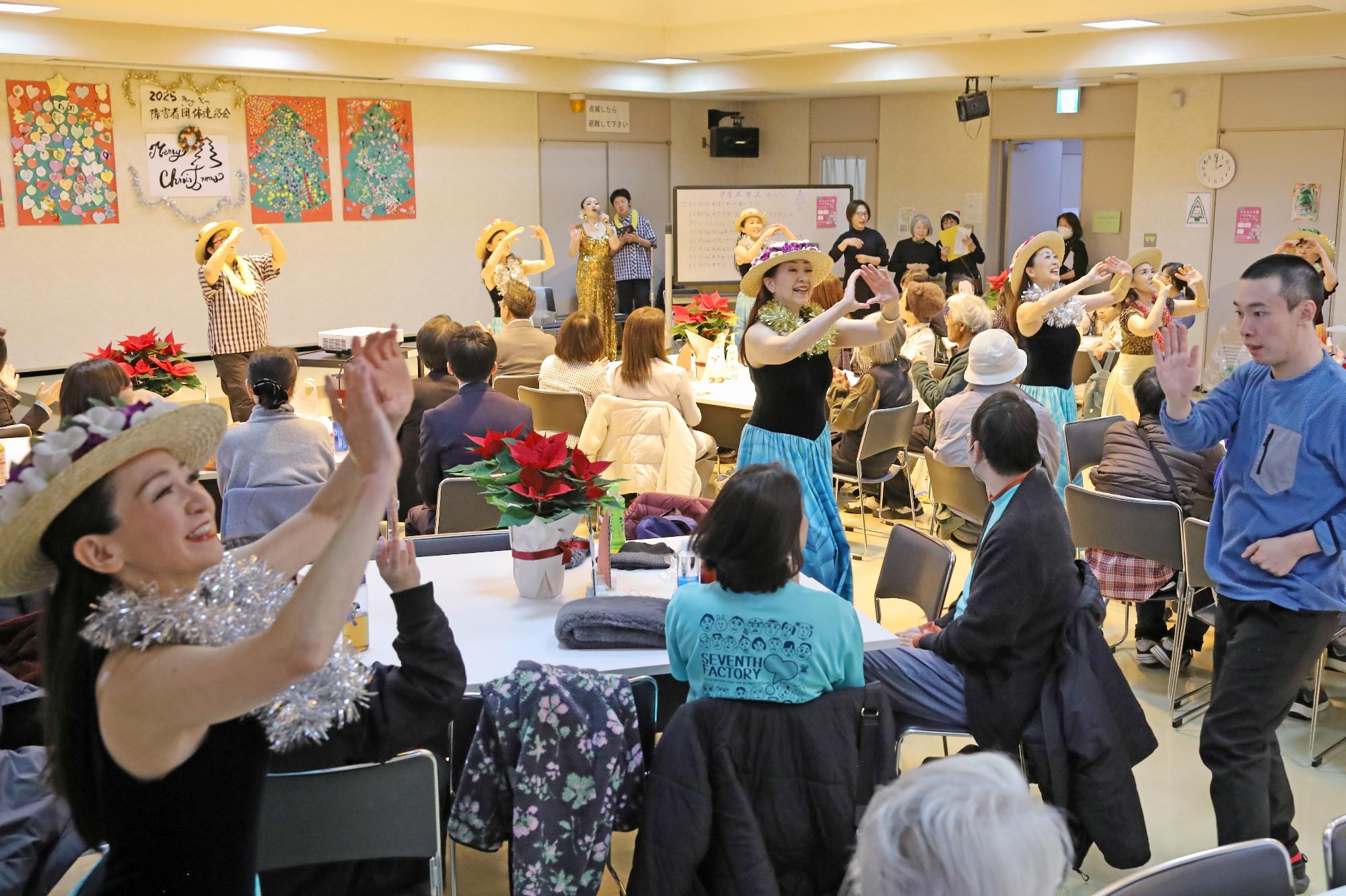 会場内でフラの衣装に身を包んだ出演者と参加者が踊りを楽しむ様子の写真