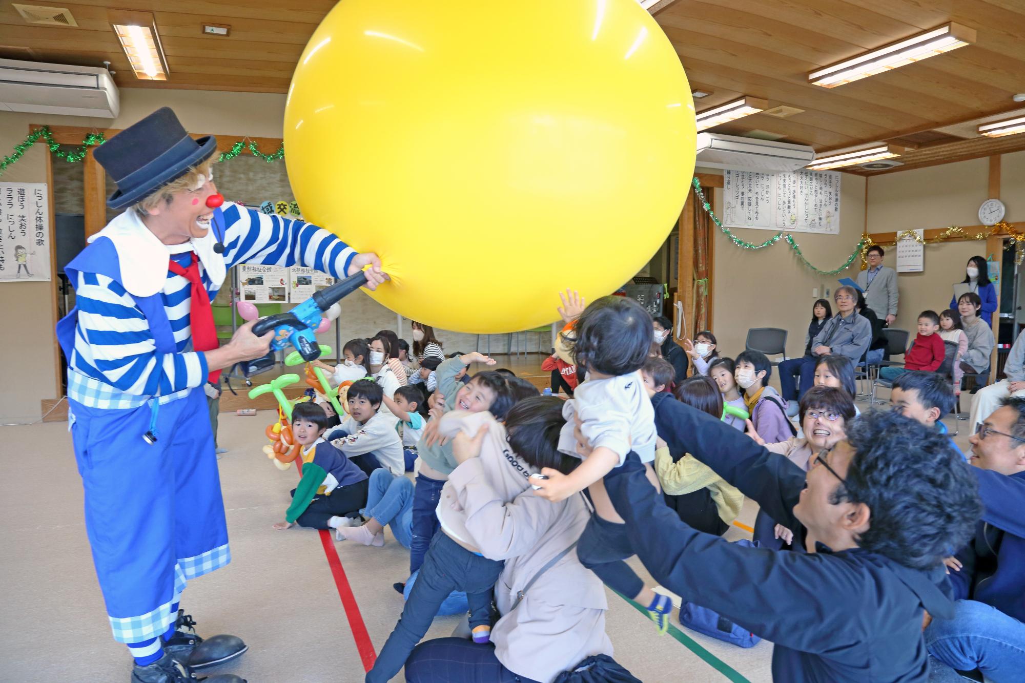 巨大な風船に空気を入れるピエロと興奮するこどもたちの写真