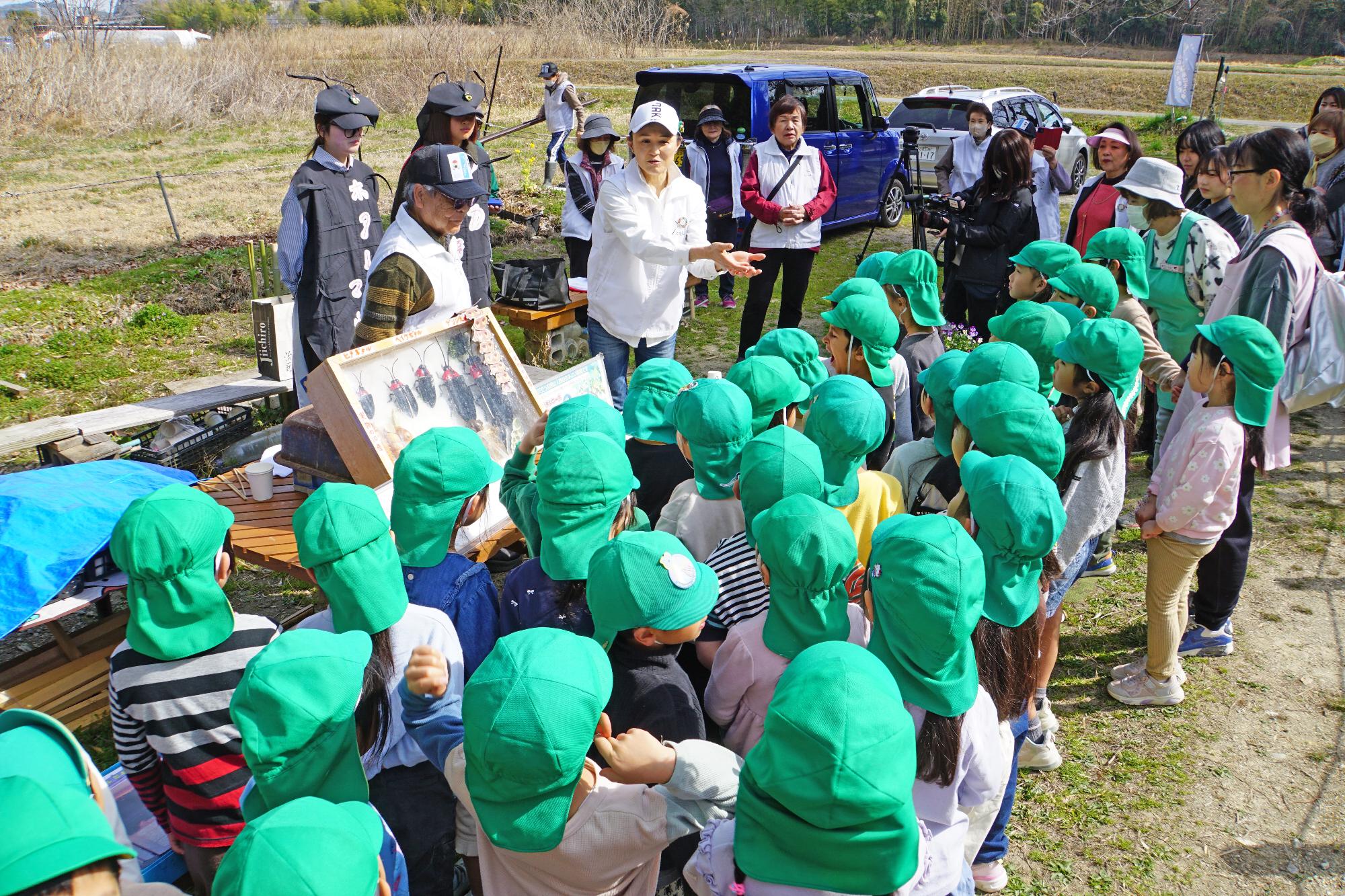 屋外で蛍の模型を見ながら蛍の解説をうける園児たちの写真