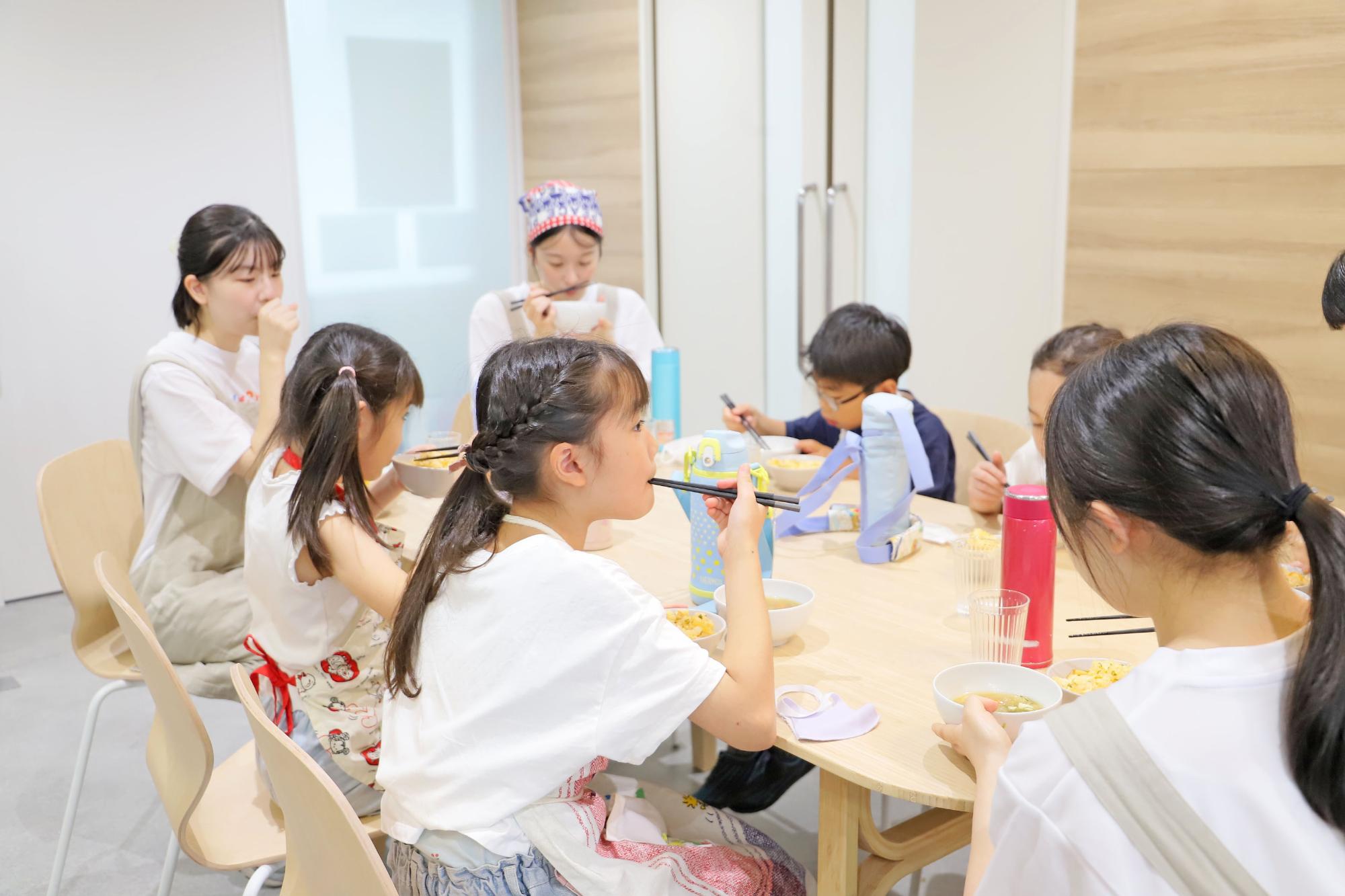 同じ机の上で出来上がった料理を食べるこどもたちの写真