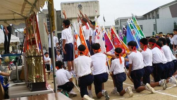 日進西中学校にてスポーツフェスティバルが開催され選手宣誓している人の周りに旗をもってしゃがんでいる人たちの写真