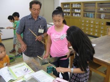 発明クラブ4年生Bクラスの子供たちが指導を受けながらのこぎりを使用して作成をしている写真